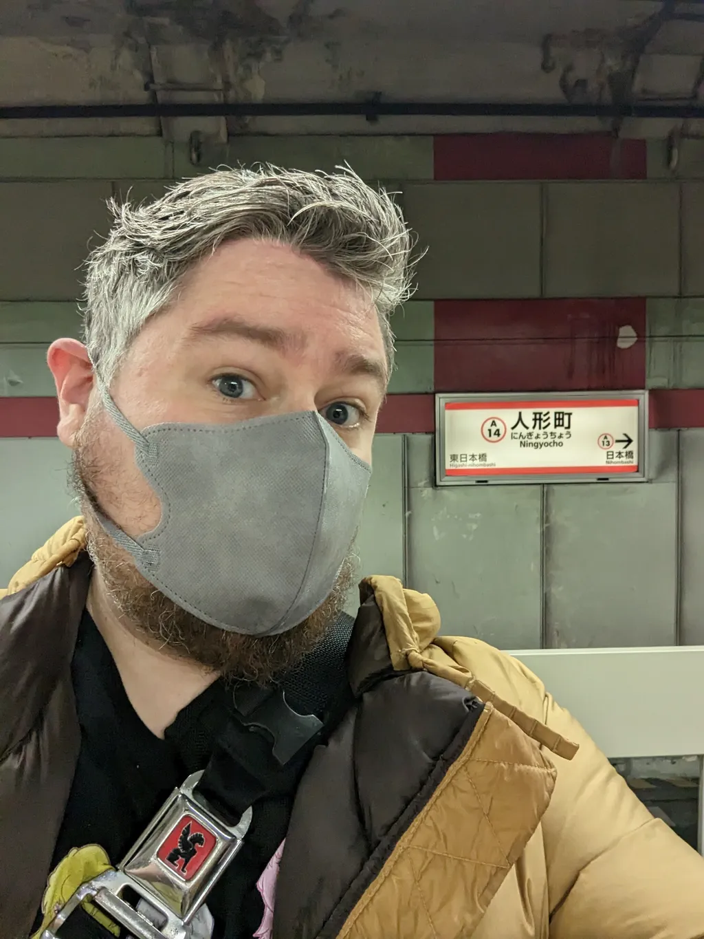 Dylan is wearing a mask and looking at the viewer. He is standing in front of a subway platform, with the 'Ningyocho' sign present in the background.